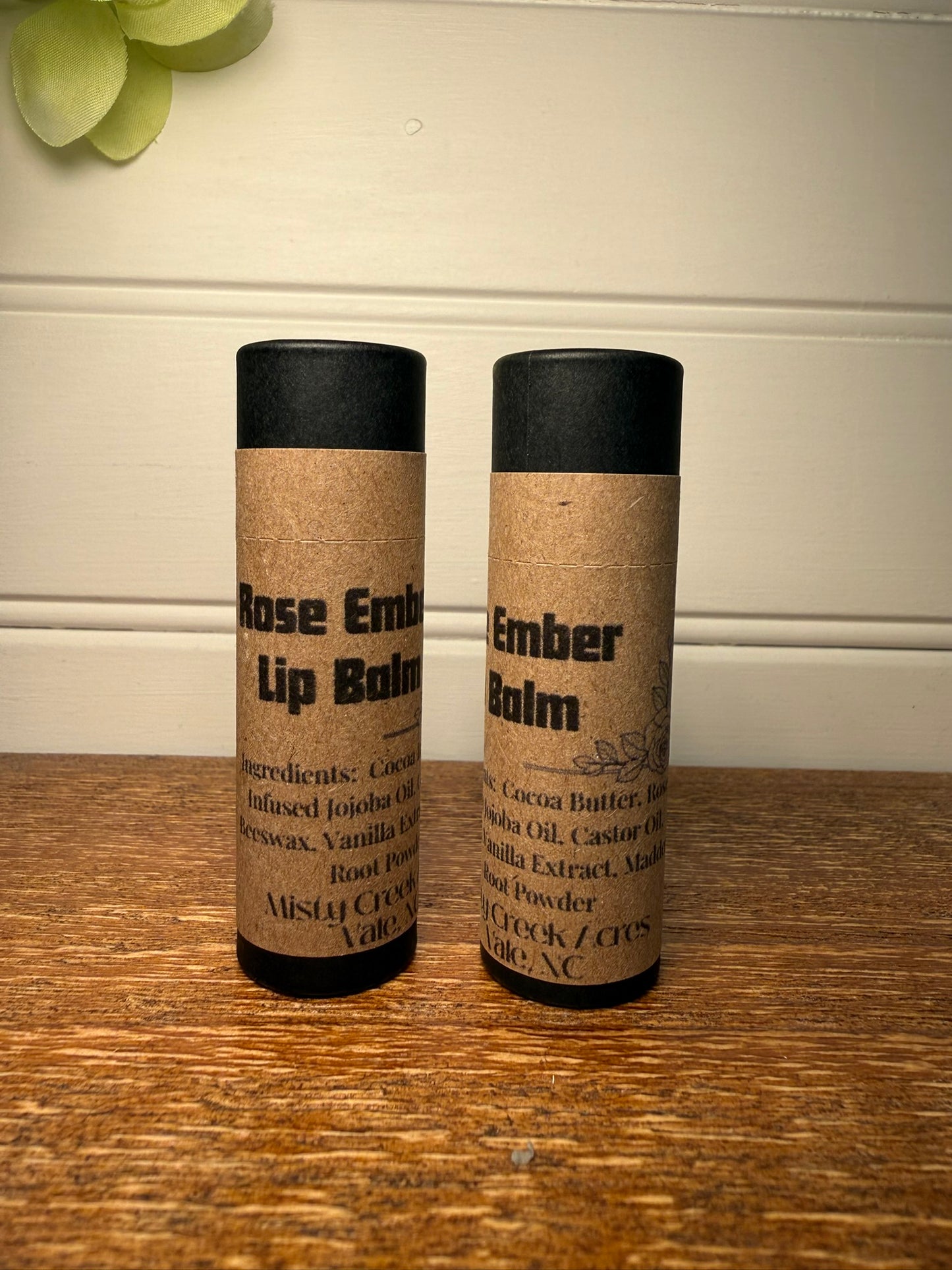 Two lip balm containers labeled 'Rose Embellished' and 'Ember Balm' on a wooden surface.