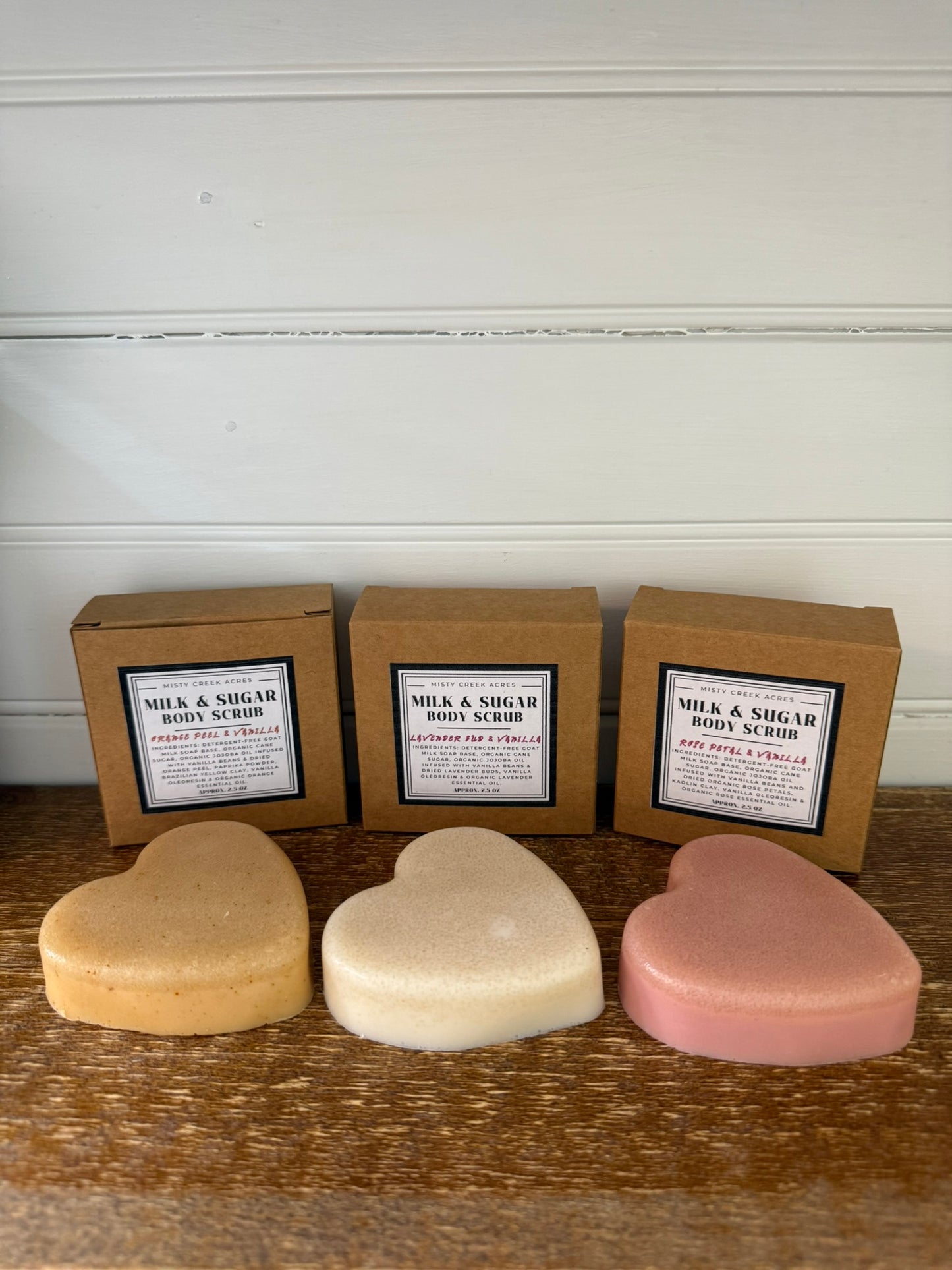 Three heart-shaped body scrubs in different colors with their packaging on a wooden surface.