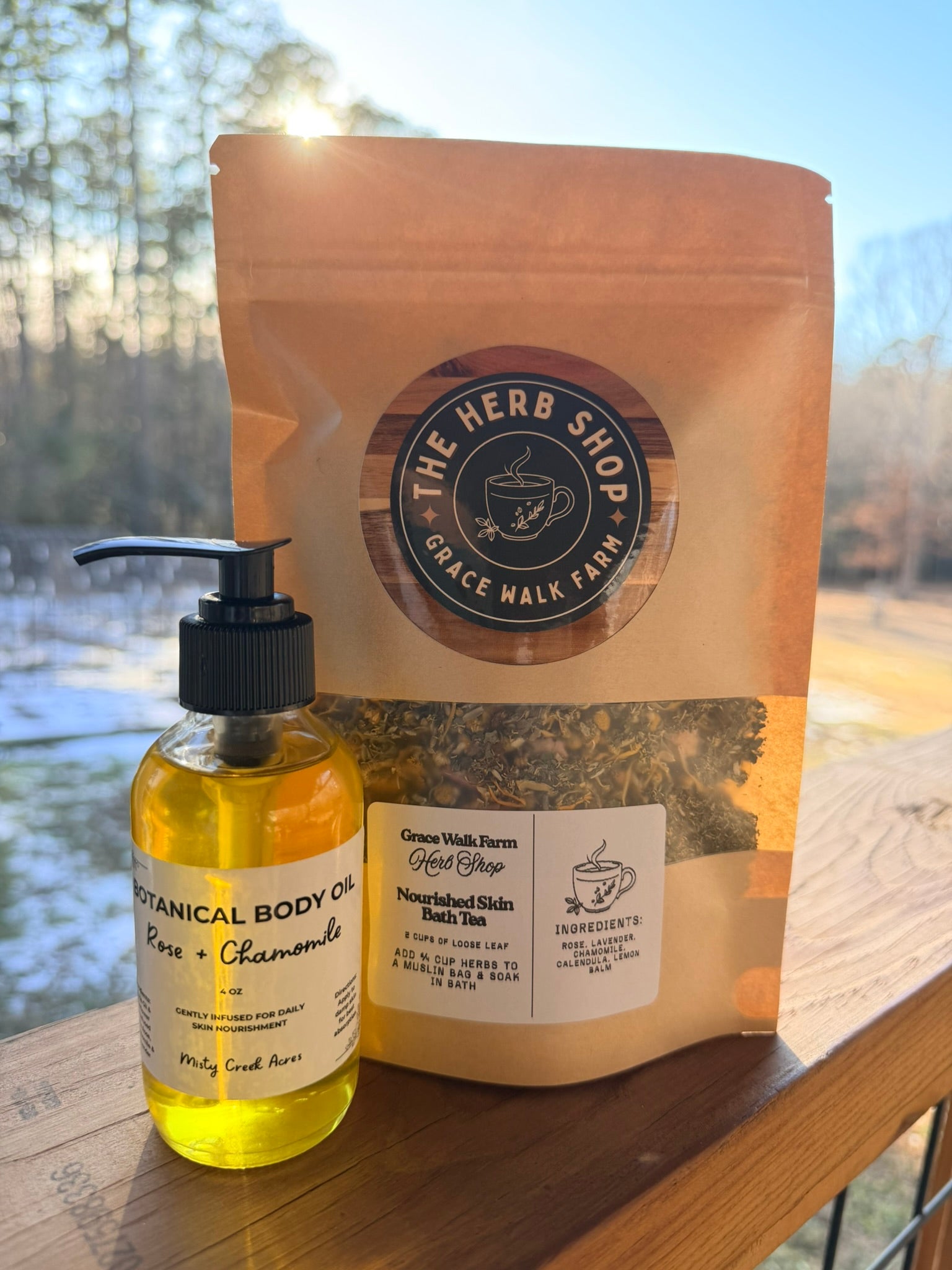 Bottle of botanical body oil and package from The Herb Shop on a wooden surface with a scenic background.