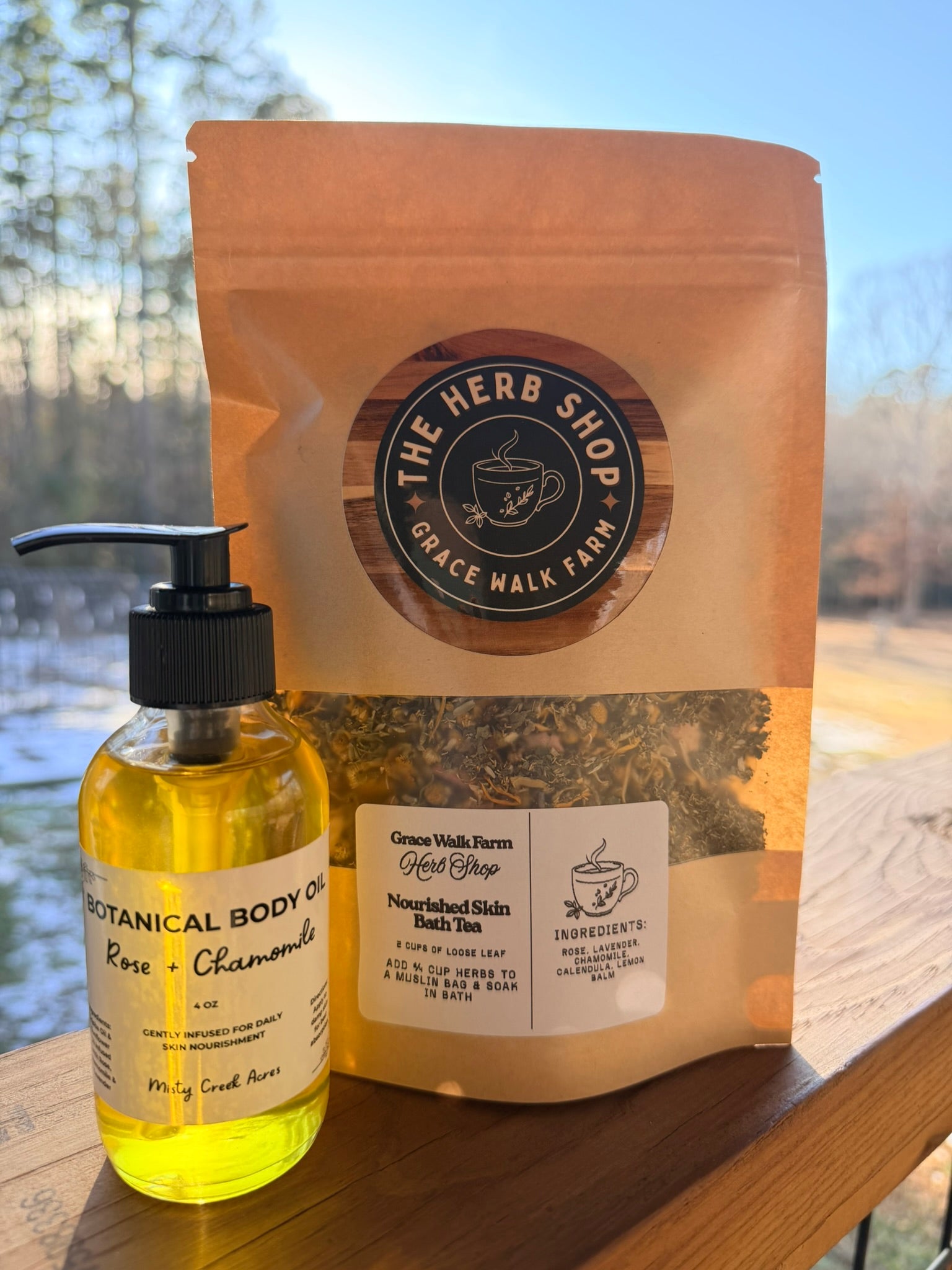 Bottle of botanical body oil and package of tea from The Herb Shop on a wooden surface with a blurred natural background.