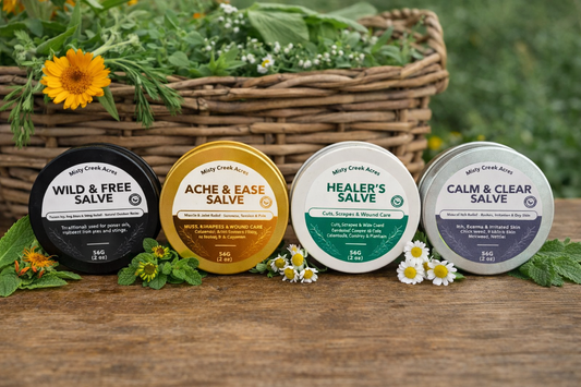 Four salve containers with labels on a wooden surface with flowers and a basket in the background.