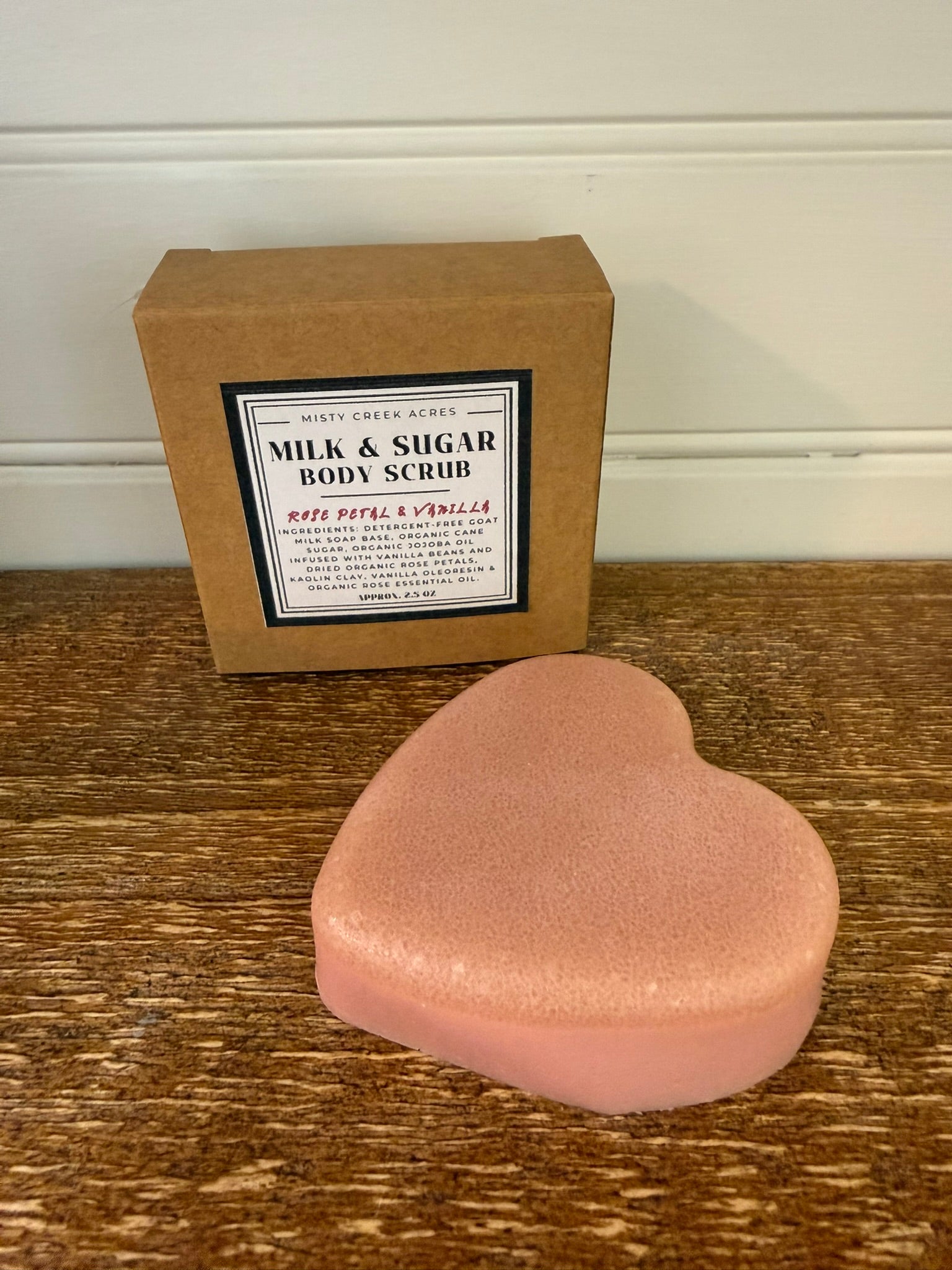 Heart-shaped pink body scrub on a wooden surface with a brown box labeled 'Milk & Sugar Body Scrub' in the background.
