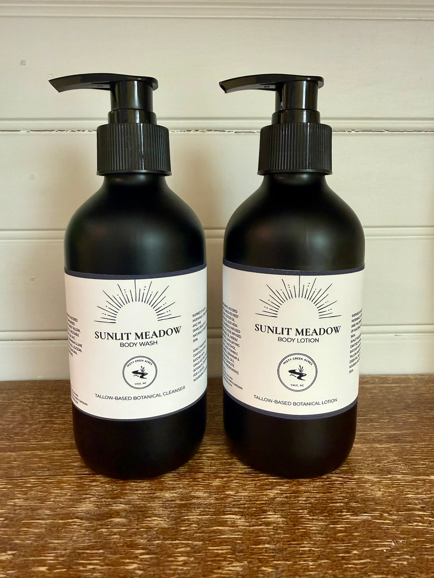 Two black bottles of Sunlit Meadow body wash and lotion on a wooden surface with a white wall background.